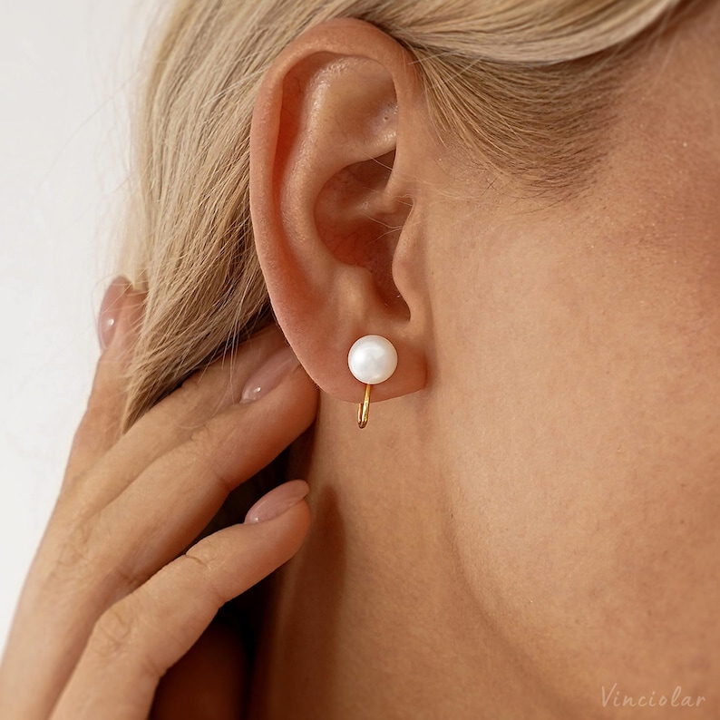May include: A close-up of a gold-colored earring with a white pearl. The earring is a small, delicate design, with the pearl positioned at the earlobe. The earring is a simple, elegant piece of jewelry.