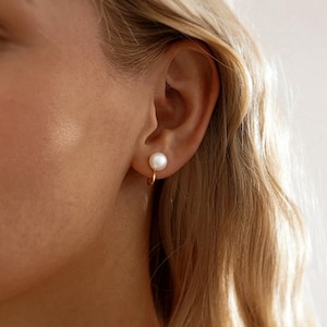 May include: A close-up of a gold hoop earring with a white pearl. The earring is attached to an ear. The person has blonde hair.