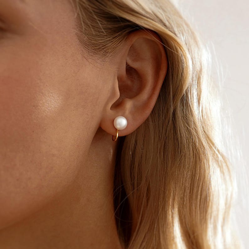 May include: A close-up of a gold hoop earring with a white pearl. The earring is attached to an ear. The person has blonde hair.