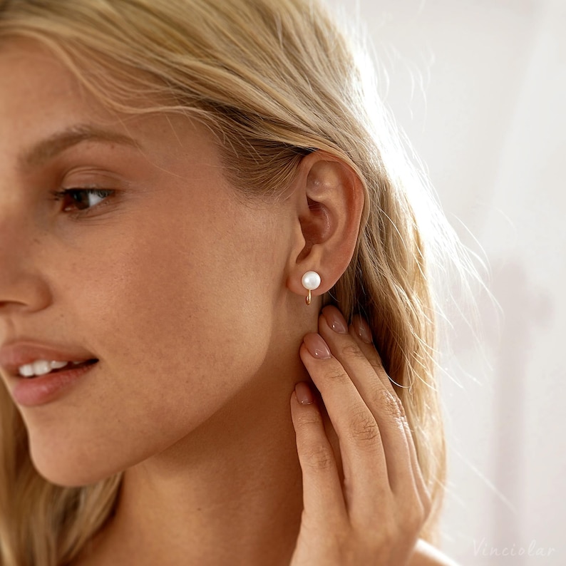 May include: A close-up of a person's ear wearing a single pearl earring. The earring features a small, round, white pearl suspended from a gold-colored hoop. The person's skin is fair, and their blonde hair is visible.