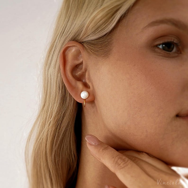 May include: A close-up of a gold-colored hoop earring with a white pearl. The earring is worn on the ear of a person with blonde hair. The earring is a simple, elegant design.