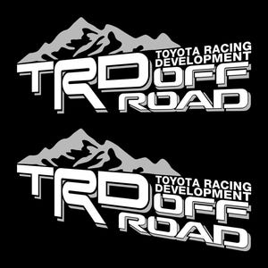 Op de afbeelding: Twee identieke witte en grijze TRD Off Road stickers op een zwarte achtergrond. De stickers tonen de tekst "TRD" boven "OFF ROAD" met "TOYOTA RACING DEVELOPMENT" ertussen. Een grijze bergketen staat boven de tekst "TRD".