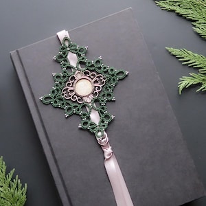 May include: A decorative bookmark in green and pink, placed on a closed black book. The bookmark has an ornate design. A lit candle, a cup of tea, and foliage are arranged on a dark surface.