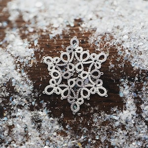 May include: A white, intricately designed snowflake ornament rests on a wooden surface, surrounded by artificial snow. The snowflake features a delicate, openwork pattern, perfect for winter holiday decor.