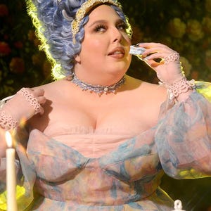 May include: A person in a light pink and blue floral dress and a blue wig, decorated with butterflies, holds a small object. The scene features lit candles, a tea set, and pastries, creating a whimsical, fairytale atmosphere.