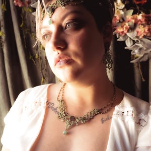 May include: An ornate, olive-green necklace, matching earrings, and a headpiece with a central gemstone. The jewellery is delicate and detailed, with a vintage aesthetic. The woman is wearing a white top with lace trim.