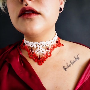 May include: A white and red lace choker necklace with dangling red beads. The necklace is worn against a dark background and a red satin top. The tattoo on the shoulder reads "thicker than dust."