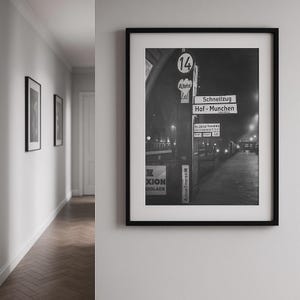Könnte beinhalten: Schwarz-weiß gerahmte Fotografie einer Bahnhofsszene. Schilder mit der Aufschrift "Schnellzug Hof-Munchen" und "XION XOLADE". Das Foto wird an einer weißen Wand mit einem Flur und weiteren gerahmten Bildern angezeigt.