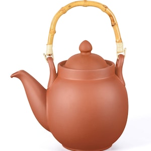 Könnte beinhalten: Eine terrakottafarbene Teekanne mit einem Bambusgriff. Die Teekanne hat einen abgerundeten Körper, einen gebogenen Ausguss und einen Deckel mit einem kleinen Knopf. Der gebogene Bambusgriff ist mit Metallbeschlägen an der Kanne befestigt.