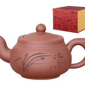 Könnte beinhalten: Eine rotbraune Keramik-Teekanne mit gebogenem Ausguss und Griff. Die Teekanne hat einen Deckel mit einem kleinen Knopf und ist mit einem Blumendesign und chinesischen Schriftzeichen verziert. Im Hintergrund befindet sich eine rote Schachtel mit goldenen Akzenten.