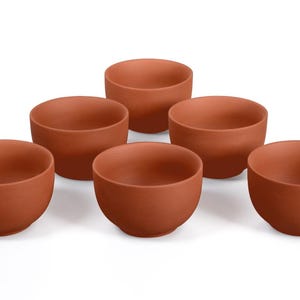Puede incluir: Seis cuencos de color terracota están dispuestos sobre una superficie blanca. Los cuencos son redondos con un acabado liso y sin esmaltar. Los cuencos son probablemente para servir alimentos o para fines decorativos.