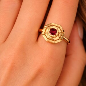 Puede incluir: Un anillo de oro con una piedra preciosa roja oscura de corte redondo engastada en el centro de un marco octogonal geométrico de varias capas. La banda de metal es lisa y pulida, mostrando un diseño estructurado y escalonado.