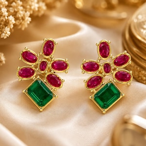 May include: A pair of gold stud earrings featuring a cluster of oval-shaped red stones arranged in a floral pattern, anchored by a single rectangular green stone at the base. The metal settings are polished gold, highlighting the vibrant contrast between the red and green gemstones in each earring.