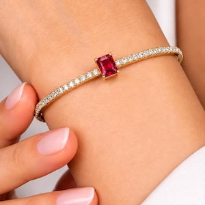 Puede incluir: Una pulsera rígida de oro con una piedra preciosa roja central de corte esmeralda, flanqueada por una hilera continua de pequeñas piedras blancas redondas y transparentes engastadas a lo largo de la fina banda de metal dorado pulido.