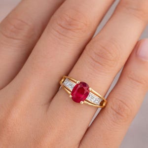 Könnte beinhalten: Ein Goldring mit einem zentralen, oval geschliffenen roten Rubin, der von vier Krappen gehalten wird. Die Ringschiene ist auf beiden Seiten in zwei parallele Goldstege geteilt, zwischen denen jeweils drei kleine, klare Diamanten im Quadrat-Schliff sitzen.