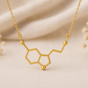 May include: A gold-toned necklace featuring a serotonin molecular structure pendant. The pendant consists of connected hexagonal and pentagonal geometric shapes with small spherical accents at the joints, suspended from a delicate gold-toned chain.