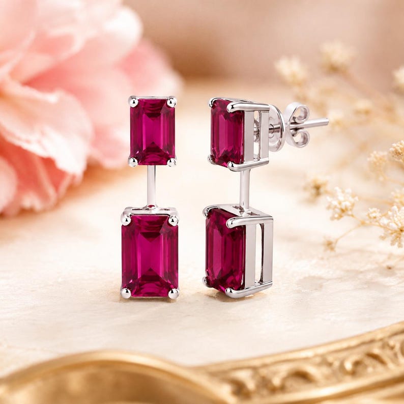 Pode incluir: Um par de brincos pendentes com duas pedras cor-de-rosa intenso em corte esmeralda, engastadas em metal prateado polido. Cada brinco consiste numa pedra superior mais pequena e uma inferior maior, ligadas por uma barra vertical com tarracha.