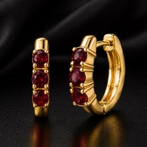 May include: A pair of gold hoop earrings, each featuring three round, deep red faceted gemstones set vertically along the front curve of the hoop. The earrings have a polished gold finish and a hinged closure mechanism visible on the side.