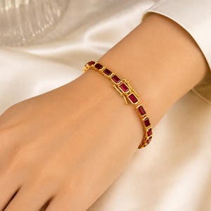 May include: A gold-toned bracelet featuring a series of rectangular, deep red gemstones set in a continuous line. The piece includes a secure, rectangular box-style clasp mechanism that matches the gold-toned metal finish of the bracelet links.