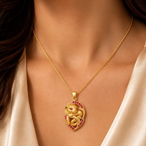 Könnte beinhalten: Eine goldene Anhängerkette mit einer skulptierten Katzenfigur mit roten Edelsteinaugen, eingefasst in einen ovalen Rahmen, der mit kleinen roten Steinen verziert ist. Der Anhänger hängt an einer feinen Goldkette und zeigt eine texturierte Oberfläche.