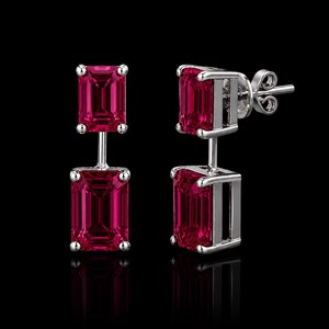 Pode incluir: Um par de brincos pendentes com duas pedras vermelho-vibrante em corte esmeralda, engastadas em metal prateado polido. Cada brinco consiste numa pedra superior menor e uma inferior maior, ligadas por uma barra vertical com fecho de pino.