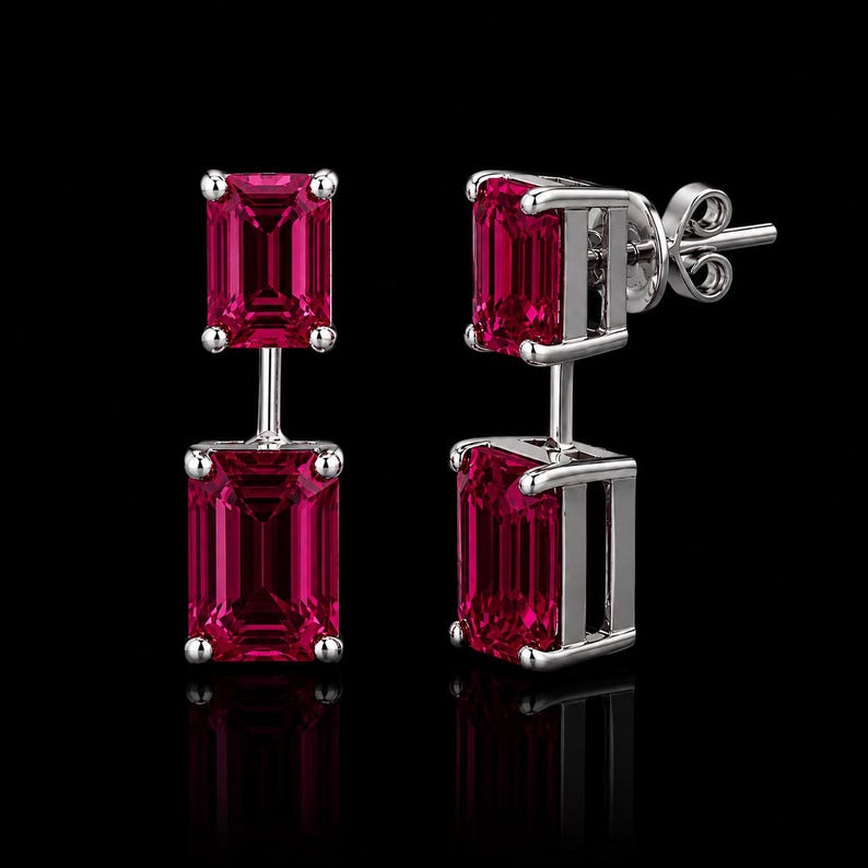 Pode incluir: Um par de brincos pendentes com duas pedras vermelho-vibrante em corte esmeralda, engastadas em metal prateado polido. Cada brinco consiste numa pedra superior menor e uma inferior maior, ligadas por uma barra vertical com fecho de pino.