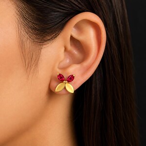 May include: A gold stud earring shaped like a butterfly, featuring two marquise-cut red gemstones as the upper wings and two smooth, solid gold petals as the lower wings, secured to the earlobe with a post and backing mechanism.