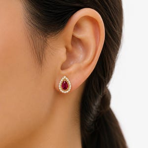 May include: A gold stud earring featuring a central pear-shaped red gemstone surrounded by a halo of small, clear, round-cut white stones. The earring is set in a polished gold metal frame and is worn on the earlobe, showcasing a classic teardrop design.