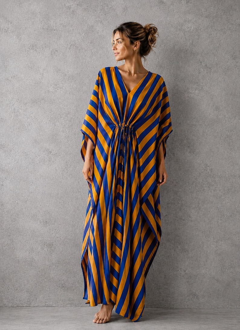 May include: A floor-length kaftan dress featuring a bold diagonal stripe pattern in vibrant royal blue and golden yellow. The garment has a deep V-neckline, wide flowing sleeves, and a central drawstring waist detail that creates soft vertical folds.