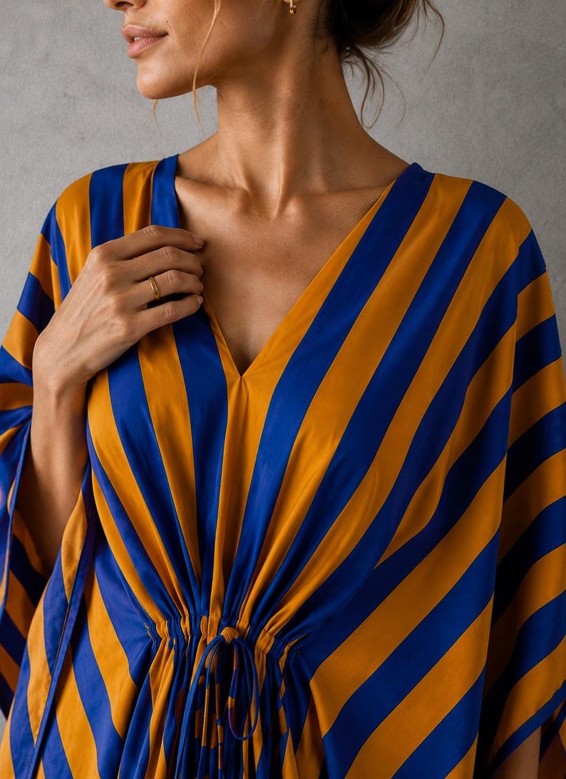 May include: A kaftan featuring a bold pattern of alternating vertical stripes in vibrant mustard yellow and royal blue. The garment has a deep V-neckline and a gathered waist detail with thin fabric ties centered at the front, creating a draped effect.