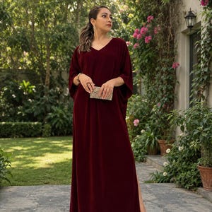 May include: A floor-length, deep burgundy velvet kaftan dress featuring a V-neckline, wide kimono-style sleeves, and a high side slit. The garment is paired with a small, rectangular, metallic gold clutch bag and strappy gold sandals.