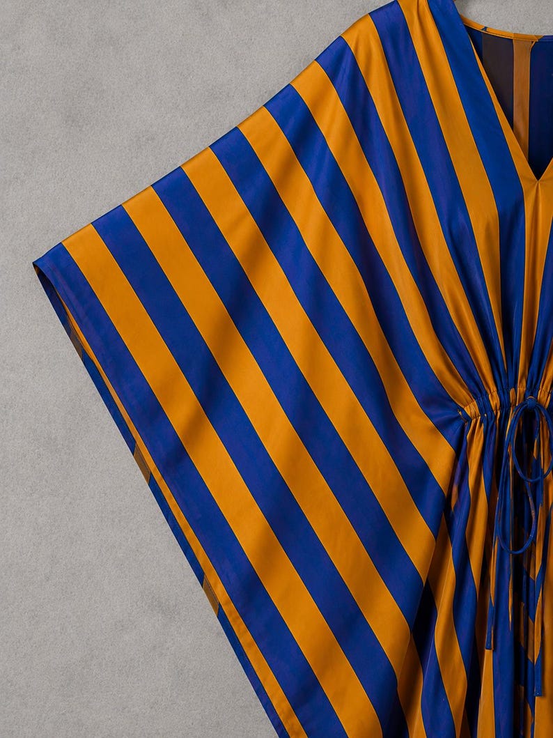 May include: A kaftan featuring a bold vertical stripe pattern in vibrant royal blue and golden yellow. The garment has a V-neckline, wide sleeves, and a drawstring waist detail with matching fabric ties that create a gathered effect at the center.