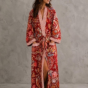May include: A long-length robe in a deep red hue featuring a vibrant, multi-colored floral and paisley pattern. The garment includes long sleeves, two front patch pockets, and a vertical white and red striped trim along the front opening.