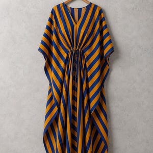 May include: A kaftan dress featuring a bold pattern of vertical and diagonal stripes in deep blue and mustard yellow. The garment has a V-neckline, wide flowing sleeves, and a central drawstring waist tie that gathers the fabric at the midsection.