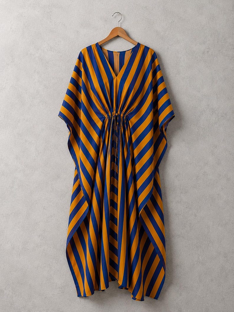 May include: A kaftan dress featuring a bold pattern of vertical and diagonal stripes in deep blue and mustard yellow. The garment has a V-neckline, wide flowing sleeves, and a central drawstring waist tie that gathers the fabric at the midsection.
