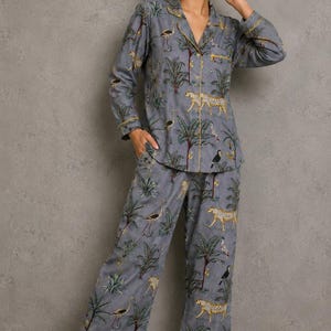 May include: A two-piece pajama set in a grey fabric featuring a repeating print of leopards, tropical birds, and palm trees. The long-sleeved button-down shirt has a notched collar and yellow piping, while the matching pants have a straight-leg cut.