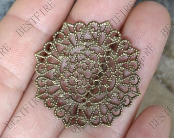 May include: A gold metal filigree charm with a circular design. The charm has a delicate, intricate pattern with a raised center.