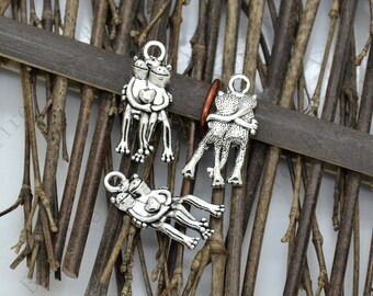 Könnte beinhalten: Drei silberne Anhänger mit Fröschen in einer Umarmung. Die Anhänger sind mit Herzen verziert und haben eine Öse zum Anbringen an einer Halskette oder einem Armband.