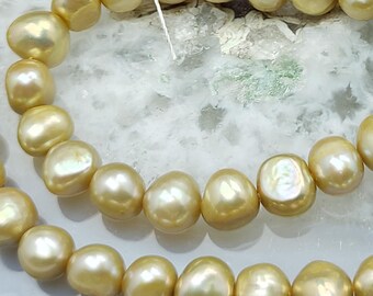 May include: Close-up of strands of golden-colored pearls. The pearls have an irregular, organic shape and a lustrous surface. The pearls are strung together with a thin, white string. The background is a light-colored, textured surface.