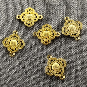 May include: Five gold-colored metal charms with a decorative design. Each charm has a circular center with a stylized pattern. The charms are arranged on a gray fabric background.