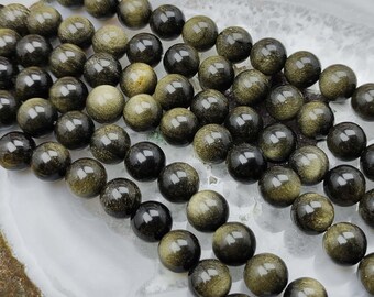 May include: A collection of round, polished beads featuring a dark black base with shimmering gold-toned inclusions. The beads are arranged in multiple parallel strands, showcasing a reflective, metallic sheen across their smooth, spherical surfaces.
