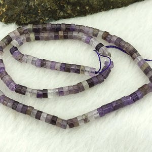 May include: Strands of amethyst gemstone beads. The beads are small, square-shaped, and vary in shades of purple, lavender, and clear. The beads are strung on a purple cord, and the background is a soft, white fabric.