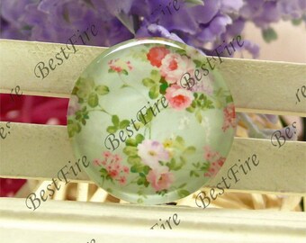 Puede incluir: Un cabujón redondo de vidrio con un patrón floral. El diseño presenta flores rosas, blancas y verdes sobre un fondo azul claro.