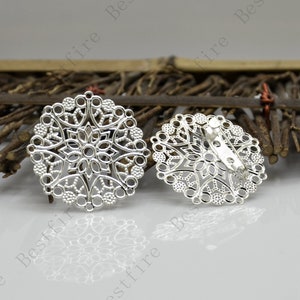 Op de afbeelding: Twee zilverkleurige filigraan broches. Elke broche heeft een cirkelvorm met een ingewikkeld opengewerkt ontwerp met bloemen- en geometrische patronen. Eén broche heeft een speld aan de achterkant. De broches worden op een lichtgekleurd oppervlak gepresenteerd.