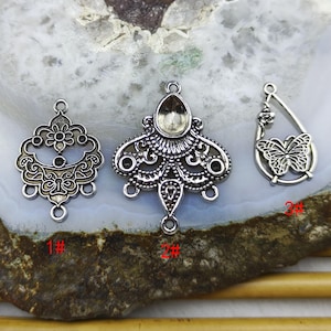 May include: Three silver-toned metal jewelry pendants. The pendants include a floral design, an ornate teardrop shape with a setting, and a butterfly design. Each pendant has a loop for attaching to a chain or other jewelry component.