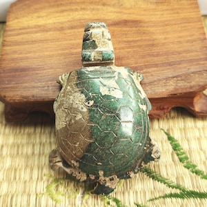 Puede incluir: Figura de tortuga tallada en verde y marrón. La tortuga tiene un caparazón texturizado con un patrón hexagonal. La cabeza y las patas también están talladas con detalles intrincados. La figura está colocada sobre una superficie de madera.