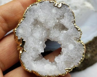 Könnte beinhalten: Ein Anhänger mit einer Geodenscheibe mit goldfarbener Umrandung und Öse. Die Geode hat ein weißes, kristallines Inneres und ein graues Äußeres. Der Anhänger ist ein Naturstein, und der Text "1#" ist sichtbar.