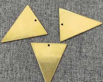 May include: Three brass triangle pendants with a small hole at the top for stringing. The pendants are a warm gold color and lie on a gray textured surface. The triangles are all the same size and shape.