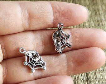 Könnte beinhalten: Zwei silberfarbene Charms mit Totenkopf- und Spinnennetz-Design. Die Charms sind perfekt, um Schmuck oder Bastelarbeiten eine gruselige Note zu verleihen.