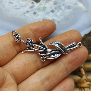 May include: A silver-toned metal jewelry connector featuring a floral design with two small rose buds and elongated leaves. The piece has three small circular loops for attachment, one at each end and one at the bottom, with a polished finish.